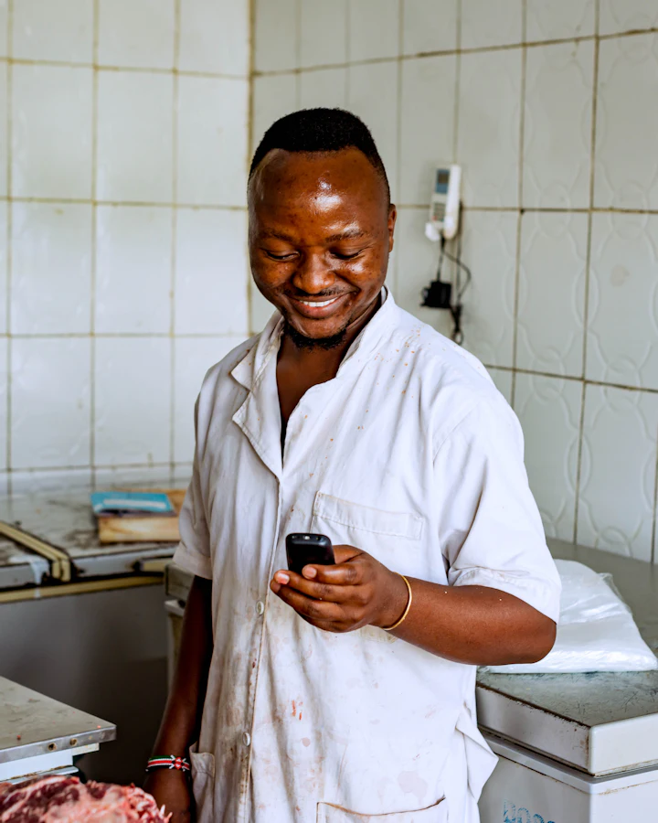 A butcher in a white coat smiles at his phone in a tiled shop.