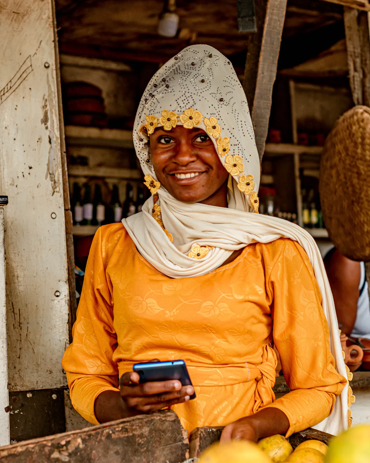 A fruit seller in a yellow top and lace headscarf smiles at her phone behind a stall of oranges.