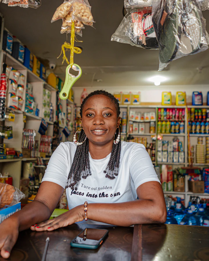 A shopkeeper leans on the counter of her mini-mart, her phone beside her.