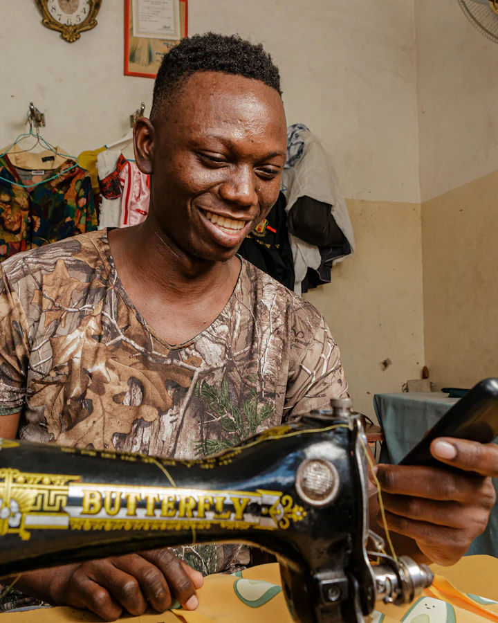 A tailor smiles at his phone, sitting at his sewing machine.